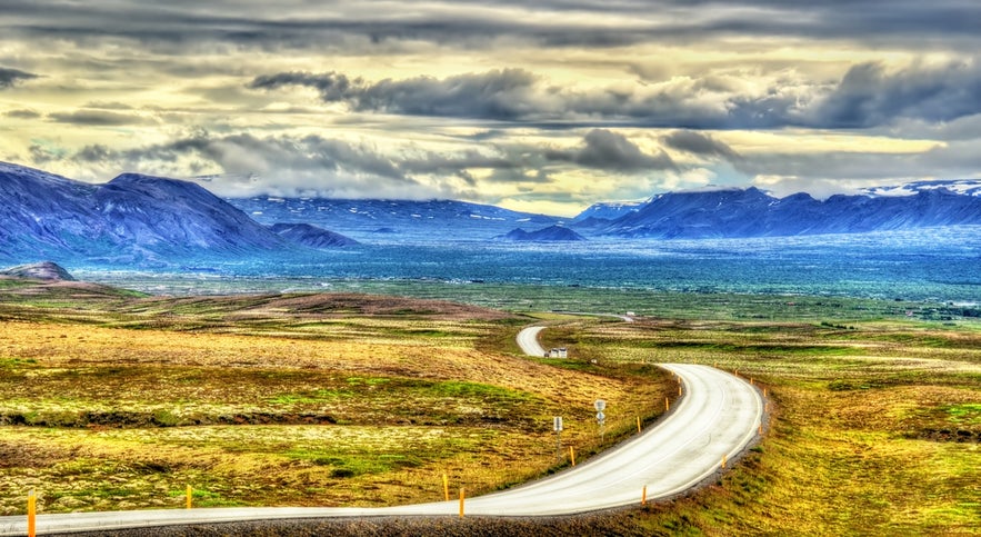 Winding road through a vast Icelandic valley with dramatic mountains and cloudy sky in the background.