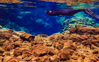 Diver exploring Silfra Fissure underwater, surrounded by volcanic rocks and crystal-clear blue water in Iceland.