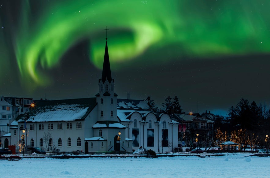 Northern lights swirl above Frikirkjan Church in Reykjavik, Iceland, with snow-covered rooftops and festive winter lights below.