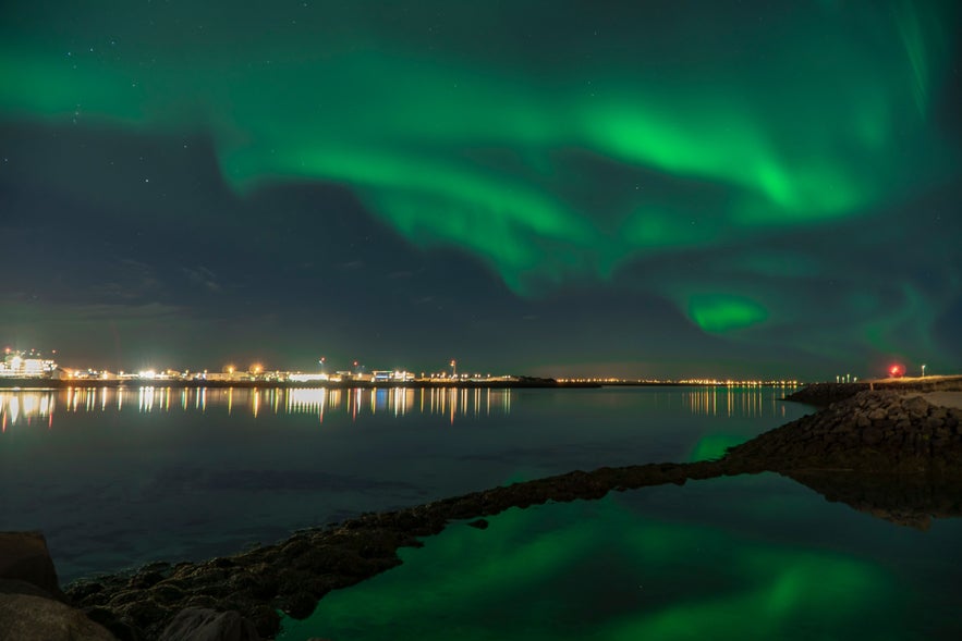 Northern lights illuminate the night sky over Reykjavik, Iceland, reflecting on the calm waters along the city shoreline.