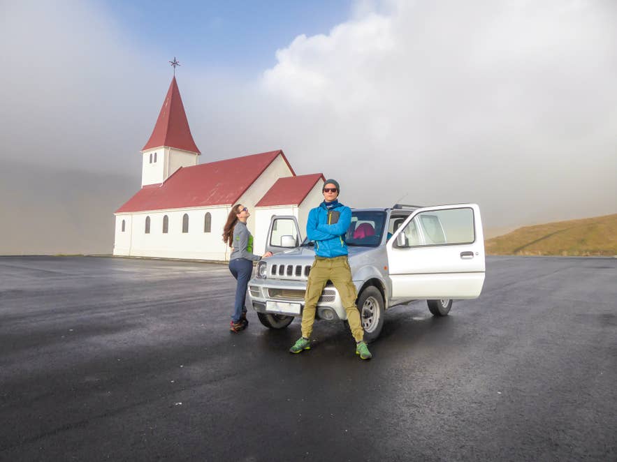 Ein Paar steht bei einem Mietwagen neben einer kleinen Kirche in Nordisland, genießt die Aussicht und amüsiert sich.