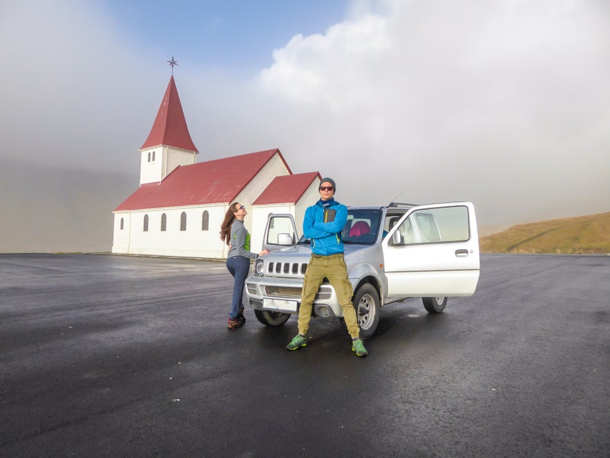 A couple stands by a rental car near a small church in North Iceland, enjoying the cloudy view and having fun.