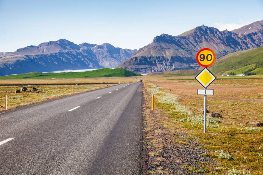 Empty paved Ring Road with a 90 km/h speed limit sign and yellow priority sign, mountains behind