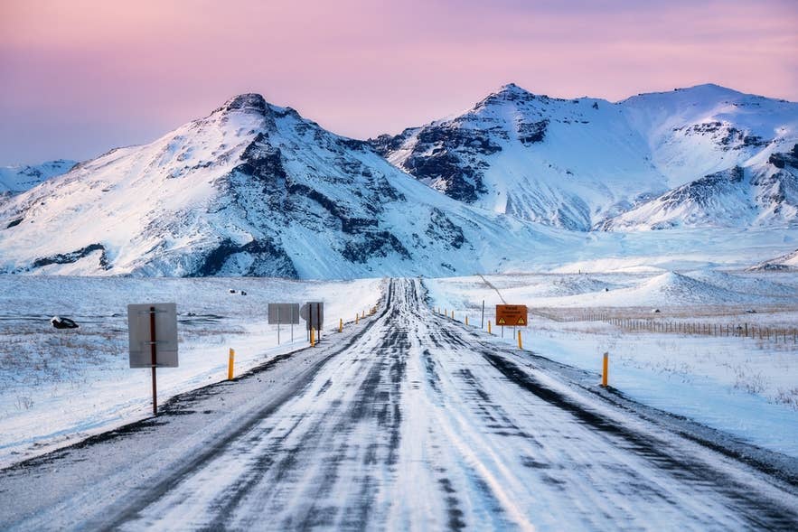 Gelbe Pfosten und Verkehrsschilder säumen die Ringstraße im Norden Islands bei Sonnenuntergang im Winter, mit schneebedeckten Bergen im Hintergrund.