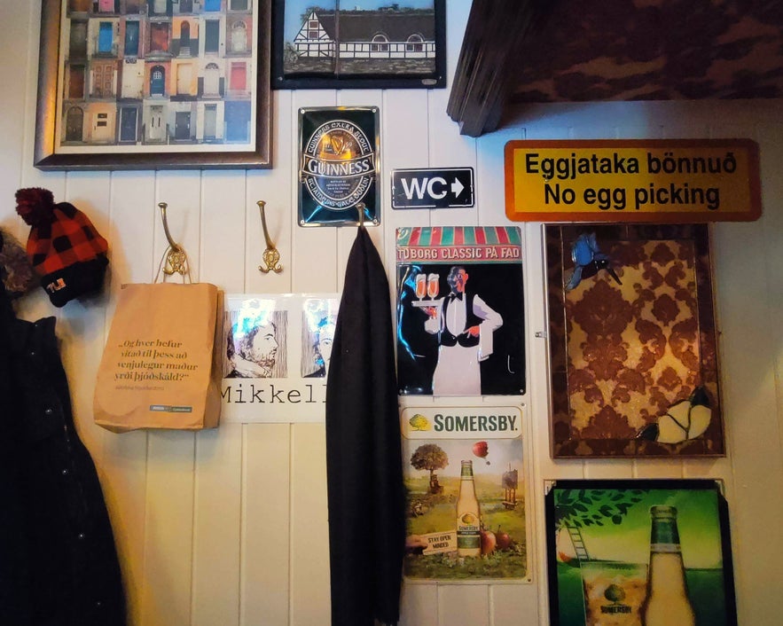 Quirky wall at Den Danske Kro in Reykjavik decorated with beer signs, vintage posters, hooks with coats and hats, and a 'No egg picking' sign.