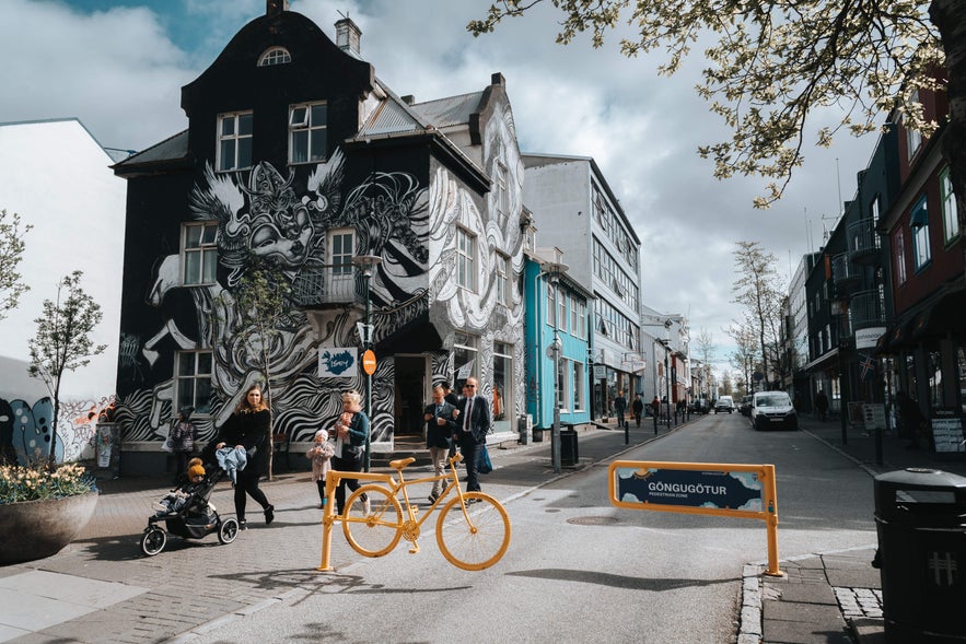 Street scene in downtown Reykjavik with people walking past a mural-covered building and colorful shops.