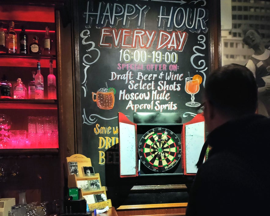 Man playing darts at The English Pub in Reykjavik, with happy hour and drink specials listed on a chalkboard.