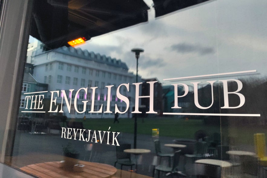 Exterior window of The English Pub in Reykjavík with logo text and reflection of Austurvollur Square and buildings.