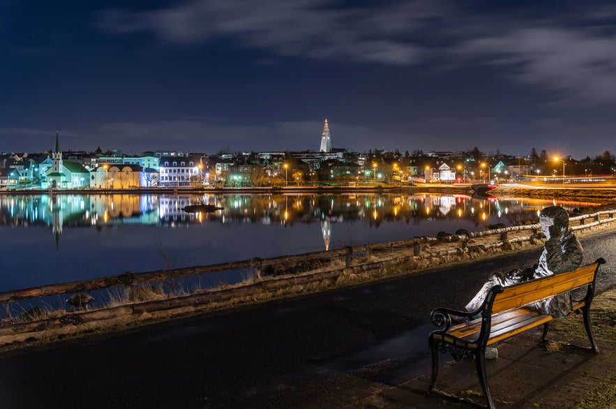 Night view of Reykjavík skyline reflected in Tjörnin pond, with Hallgrímskirkja in the distance and seated statue in foreground.