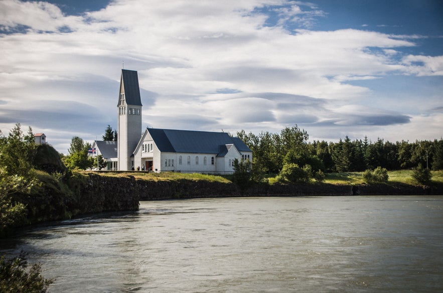 Selfosskirkja church on the banks of the Ölfusá River in South Iceland, framed by trees and clouds on a sunny summer day.