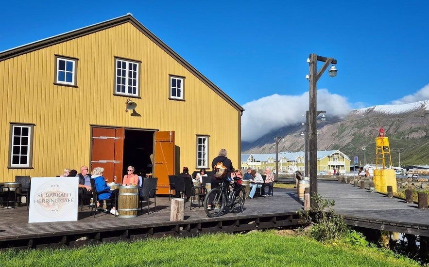 Guests dining outdoors at the Herring Café in Siglufjörður, Iceland, set in a historic yellow building by the harbor with mountain views in the background.