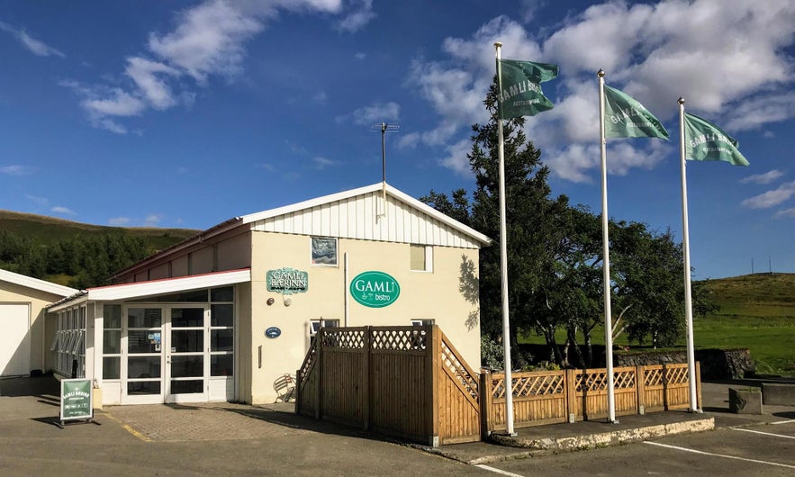 Exterior of Gamli Bærinn bistro near Lake Mývatn, Iceland, with flagpoles, a small outdoor patio, and clear blue skies above the surrounding countryside.