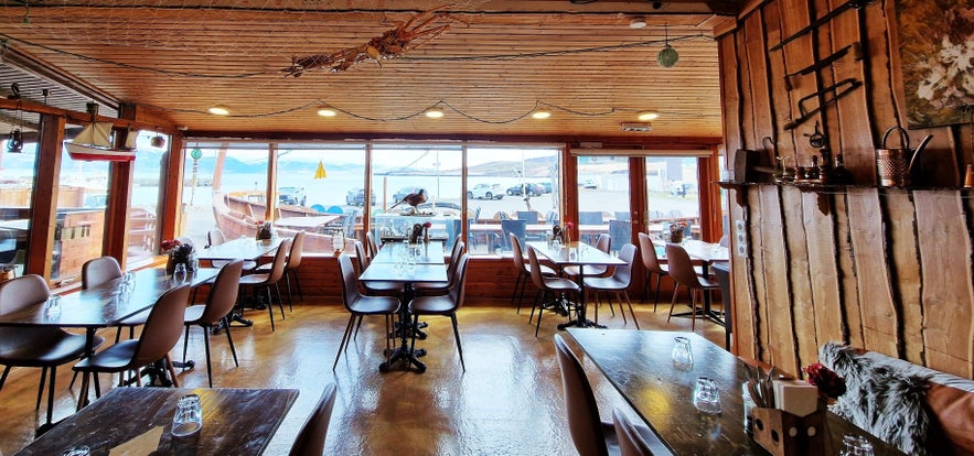Interior of Baccalá Bar in Dalvík, Iceland, with rustic wooden décor, cozy seating, and large windows offering scenic views of the harbor and surrounding mountains.