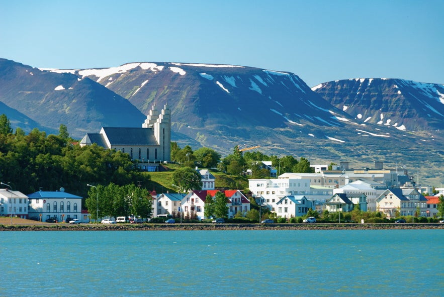 View of Akureyri, with Akureyrarkirkja church and colorful houses along the fjord, backed by snow-streaked mountains under a clear blue sky.