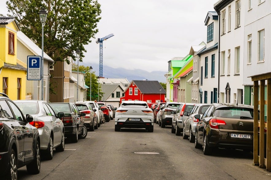 Driving in Iceland on a one-way street in downtown Reykjavik lined with colorful houses and parked cars.