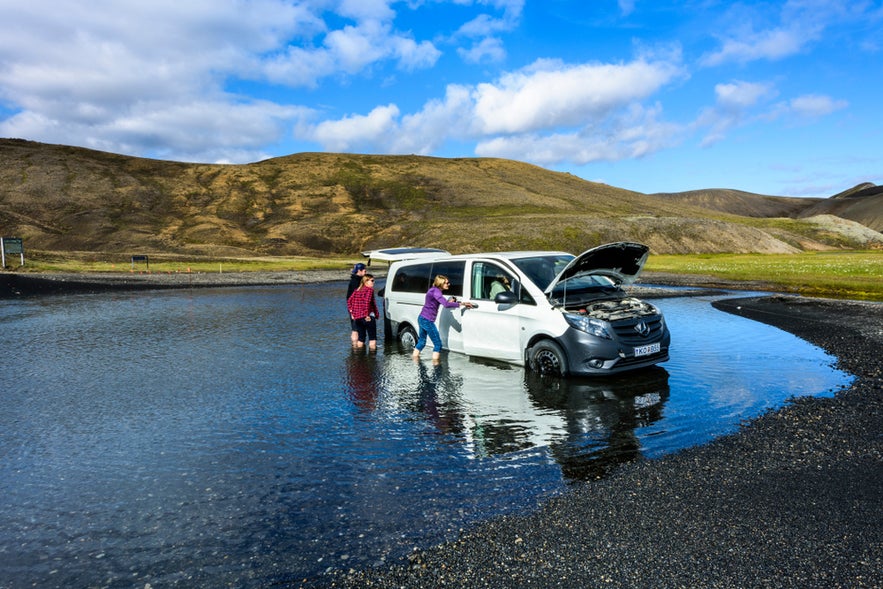 Tourists pushing a stuck rental car in a flooded area while driving in Iceland, highlighting challenging road conditions.