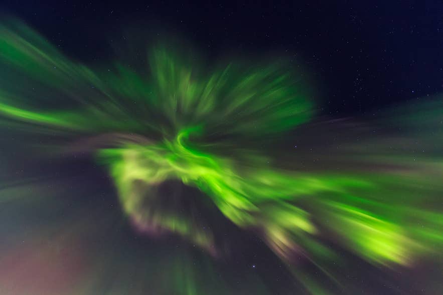 Vivaci aurore boreali verdi si avvolgono scenograficamente nel cielo notturno islandese, irradiandosi verso l’esterno con stelle appena visibili sullo sfondo.