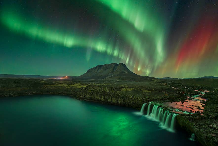 L’aurora boreale, nelle tonalità del verde e del rosso, illumina il cielo notturno sopra la cascata di Þjófafoss e una montagna impervia in Islanda, riflettendosi nel fiume sottostante.