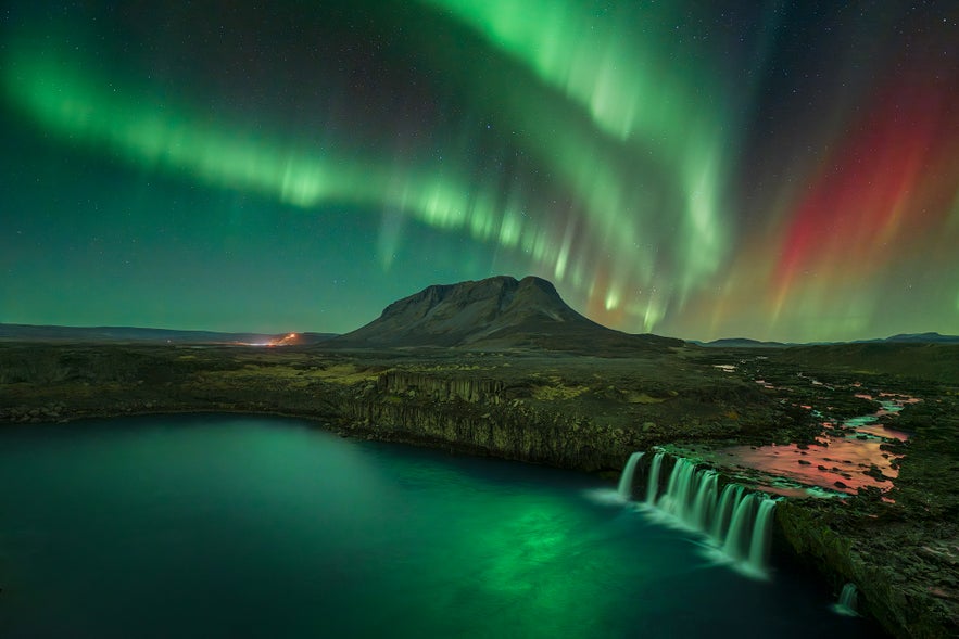 Northern lights in shades of green and red illuminate the night sky above Þjófafoss waterfall and a rugged mountain in Iceland, reflecting on the river below.