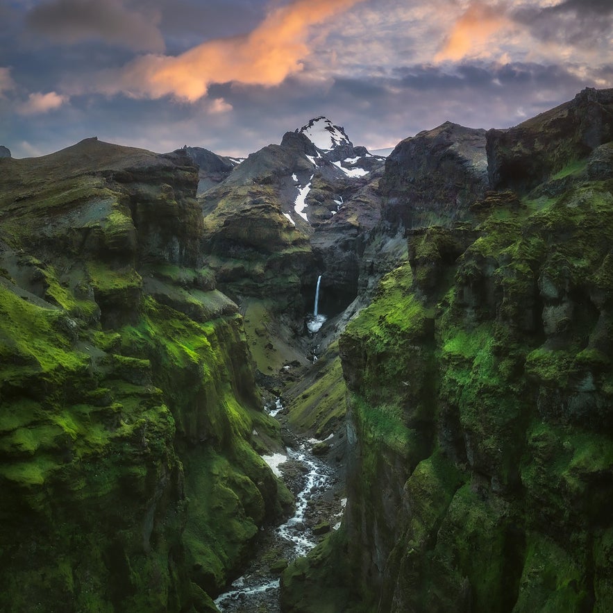 A dramatic view of Mulagljufur canyon in southeast Iceland, with steep green cliffs, a winding river, and a tall waterfall cascading from snow-capped mountains under a colorful sunset sky.