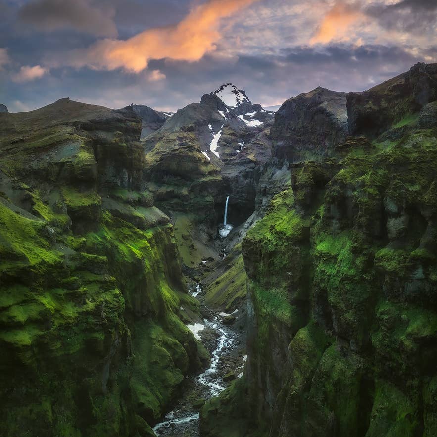 A dramatic view of Mulagljufur canyon in southeast Iceland, with steep green cliffs, a winding river, and a tall waterfall cascading from snow-capped mountains under a colorful sunset sky. A dramatic view of Mulagljufur canyon in southeast Iceland, with steep green cliffs, a winding river, and a tall waterfall cascading from snow-capped mountains under a colorful sunset sky.
