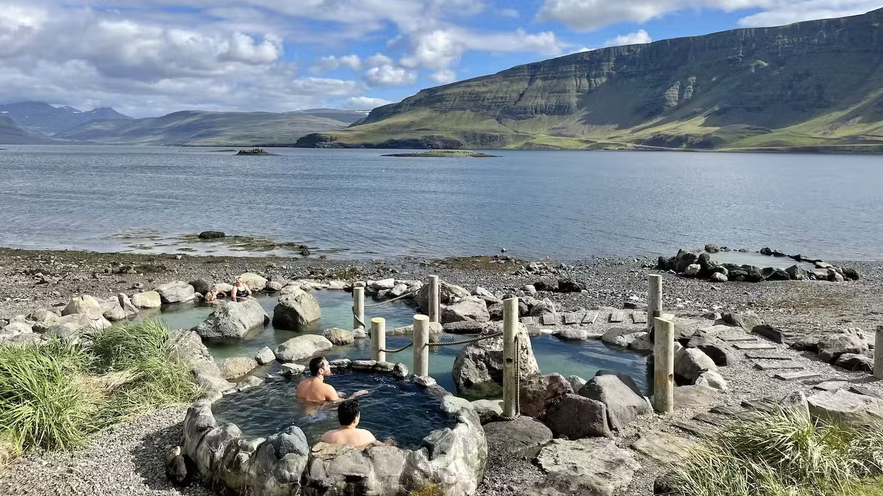 People relaxing in small stone hot pools at Hvammsvik Hot Springs, overlooking the fjord and surrounding mountains.
