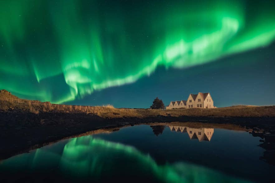 Una casa di campagna in Islanda sotto una vivida aurora boreale, riflessa magnificamente sulla superficie calma dell'acqua.