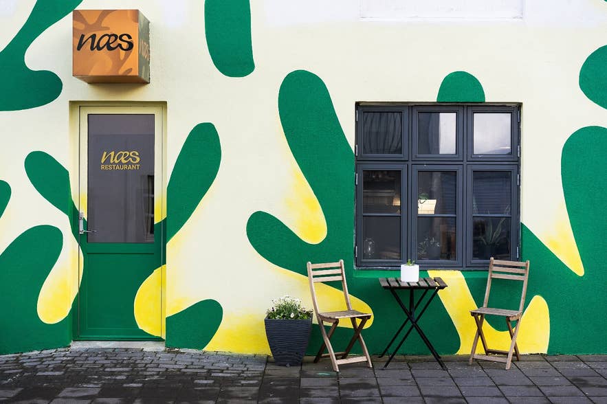 The facade of Næs restaurant in the Westman Islands, featuring a green and yellow wall with a table and chairs.