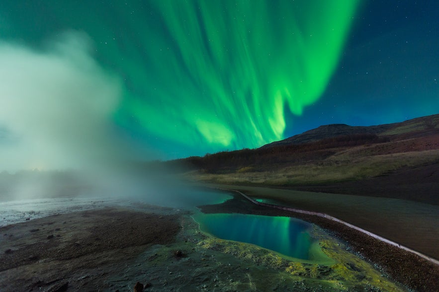 Bright green northern lights dancing in the night sky above a steaming hot spring in Iceland.