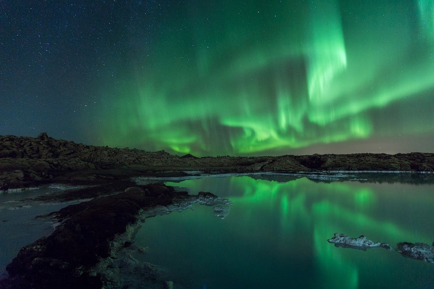 Green northern lights reflecting on the still waters of a rocky lagoon in Iceland under a starry night sky.