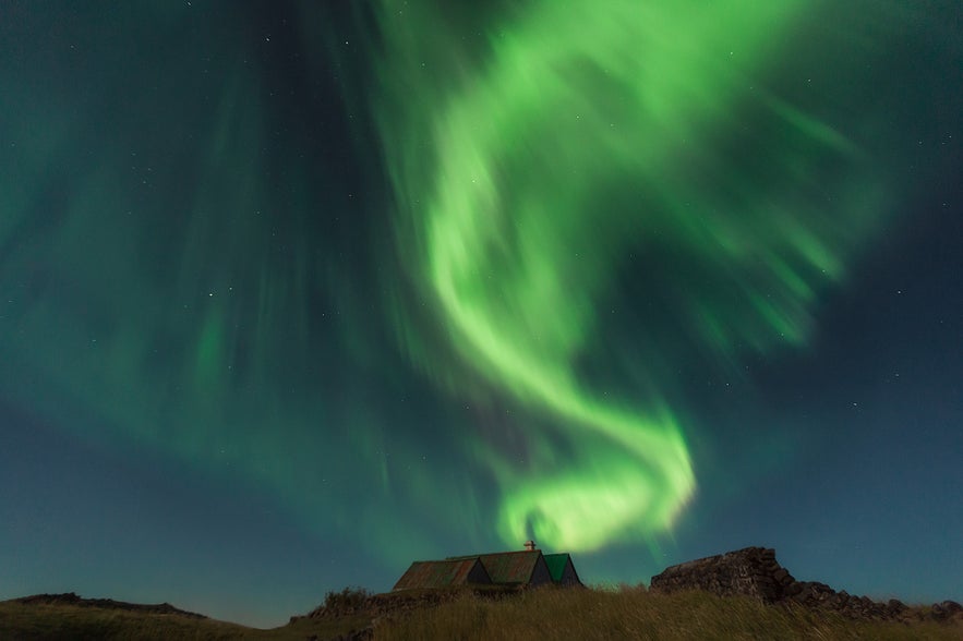 Bright green northern lights swirling above a small turf-roofed house in rural Iceland.