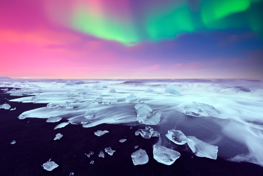 Ice chunks on Iceland’s Diamond Beach under the northern lights, with waves washing over black sand at twilight.