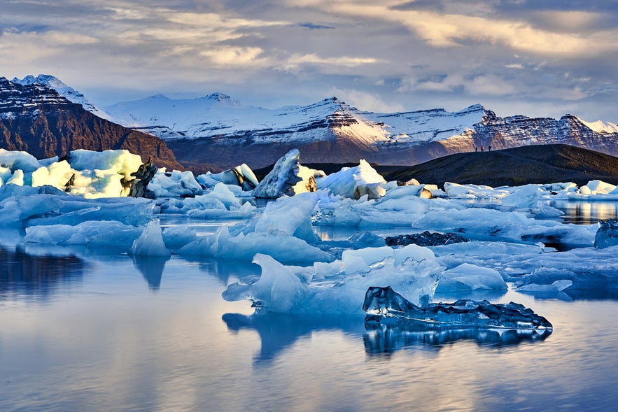 Icebergs floating in Jokulsarlon Glacier Lagoon, with snowy mountains and golden evening light in the background.