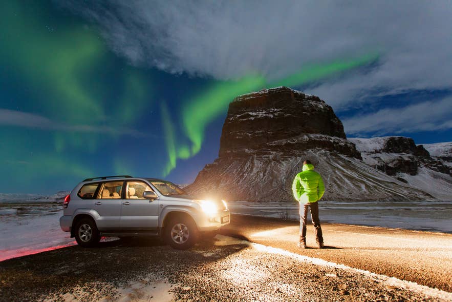 ロゥマアグヌープル山近く、ヨークルスアゥルロゥン氷河湖周辺でオーロラの下に立つ人とSUV。