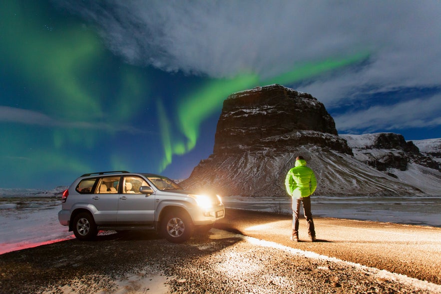 Person stands near SUV under northern lights by Lomagnupur Mountain, near Jokulsarlon Glacier Lagoon in southern Iceland.