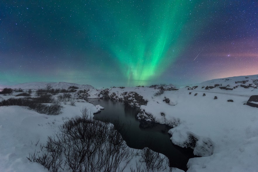 Northern lights glow above a snowy landscape and river in Thingvellir National Park, Iceland, under a starry winter sky.