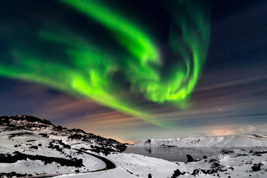 Green northern lights swirl above snowy mountains and a frozen lake at Kleifarvatn in Iceland on a clear winter night.