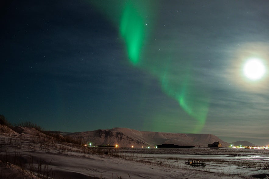 Northern lights glow above Esja Mountain near Reykjavik, with the moon illuminating the snowy winter landscape.