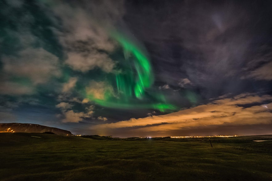 Northern lights swirl through the clouds above Ulfarsfell mountain, with Reykjavik city lights glowing on the horizon.