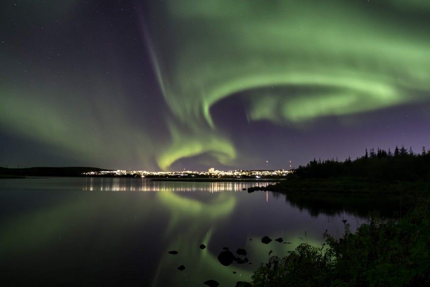 Northern lights shine above Reykjavik, reflecting on a lake in Heidmork Nature Reserve, with city lights glowing in the distance.