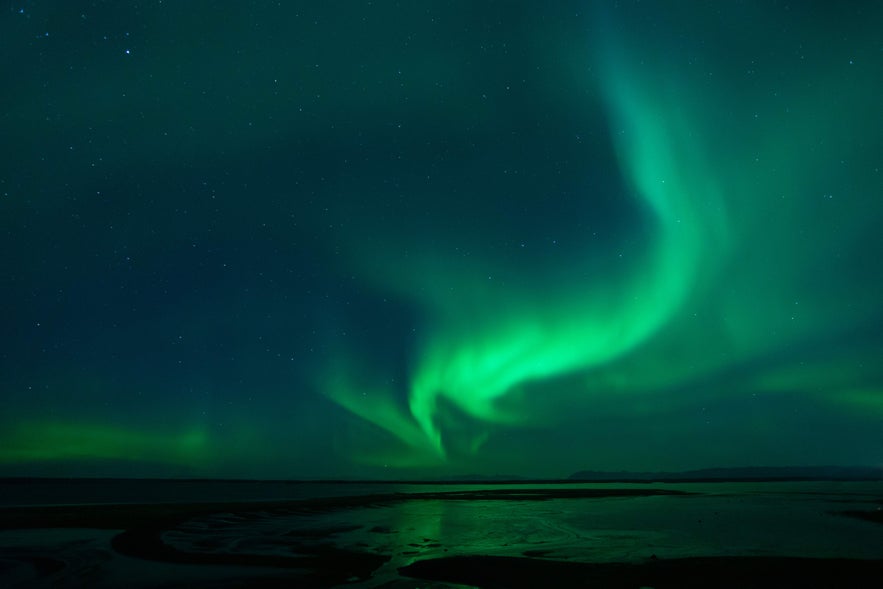 Northern lights dance in green waves over Borgarnes, reflecting on the shoreline and dark waters beneath a starry night sky.