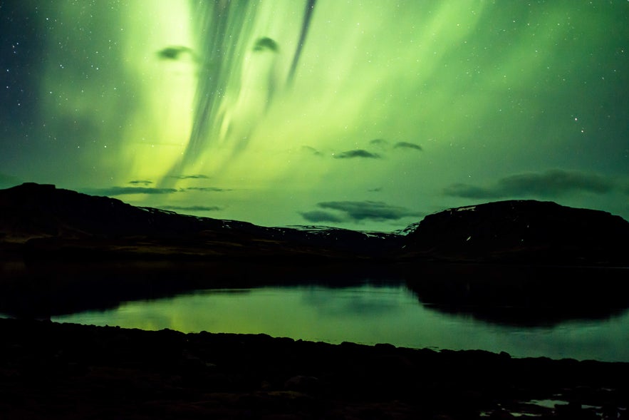 Northern lights streak across the night sky above Hvalfjordur Fjord, reflecting on the calm waters between dark mountain ridges.