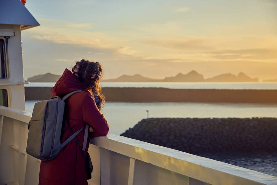 Reisender auf der Fähre bei Sonnenuntergang mit Blick auf die Westmännerinseln in Island, ruhige See und Vulkane im Hintergrund.