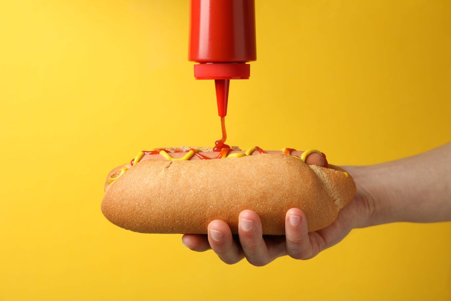 Hand holding Icelandic hot dog with mustard and ketchup, a classic snack often enjoyed by visitors in Reykjavik.