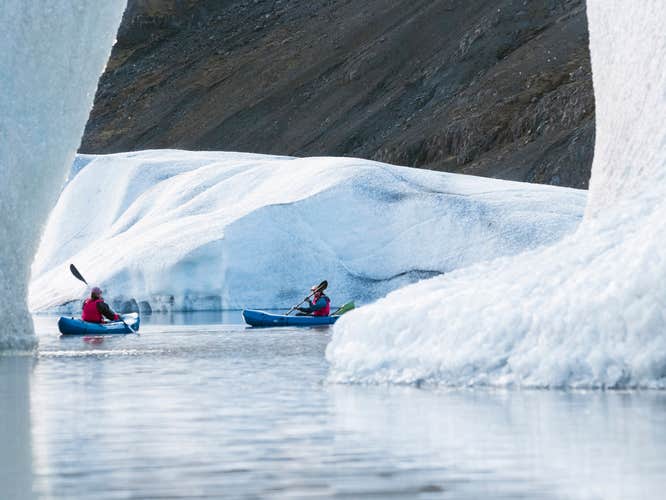 Adventurers kayaking between massive icebergs on Heinabergslon Glacier Lagoon, with striking blue ice and volcanic cliffs.