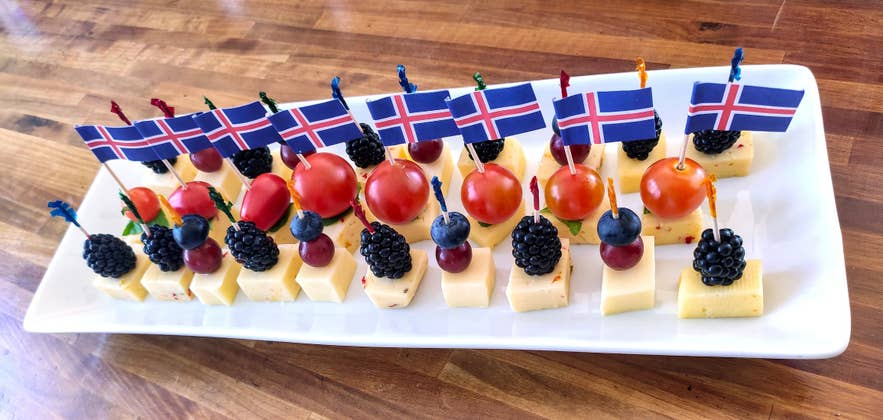 Cheese cubes topped with berries and tomatoes are served on skewers with Icelandic flags, arranged neatly on a white platter.