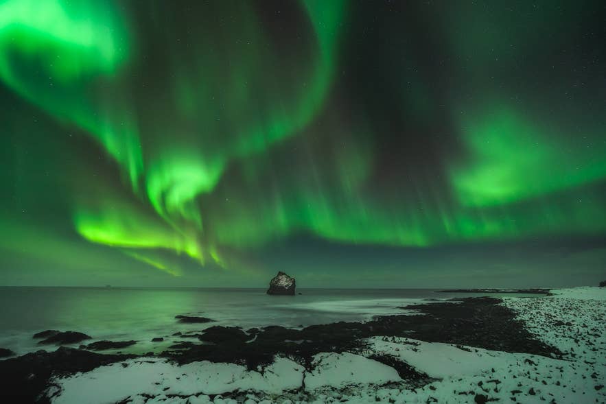Northern lights dance above a huge rock formation sitting in the sea.