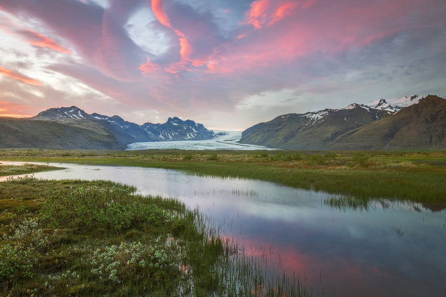 Vatnajokull Glacier flows between rugged peaks under a pink sunset sky, reflecting in a calm wetland in southeast Iceland.