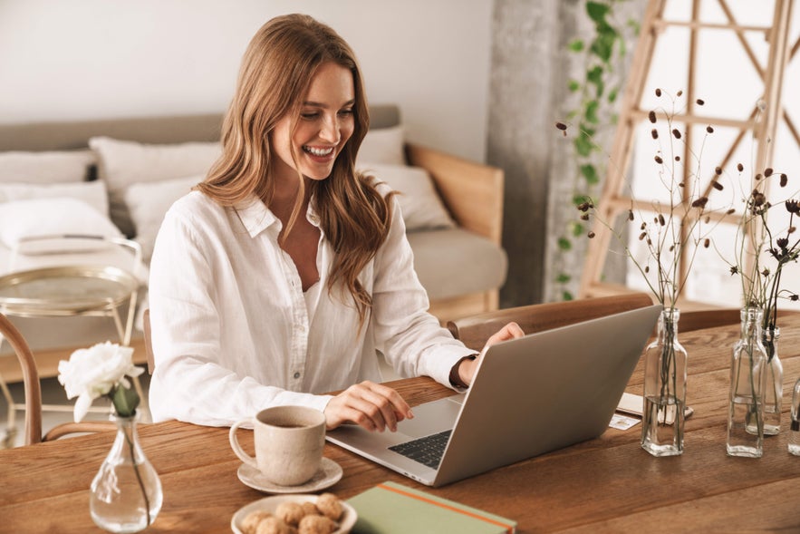 Smiling woman working on a laptop at home with coffee, researching property listings and opportunities in Iceland real estate.