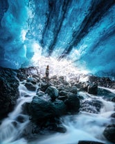 A woman stands beneath glowing blue ice in Solheimajokull cave, surrounded by rocks and rushing glacial meltwater.